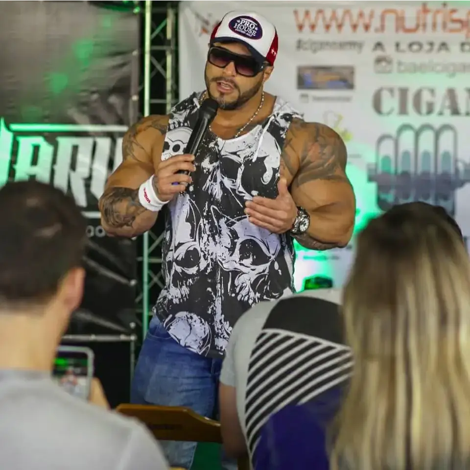 A muscular man wearing a white and red pro house cap, black sunglasses, and a sleeveless hoodie with a bold black-and-white skull pattern.
