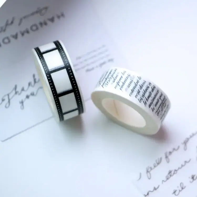 Two rolls of washi tape, one featuring a black and white film strip pattern and the other adorned with elegant cursive script in dark ink on a cream background.