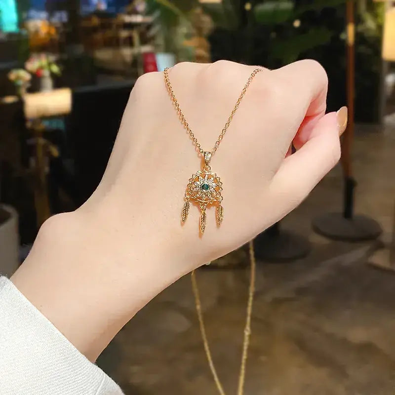 Gold-colored dreamcatcher pendant necklace with a central green gemstone and dangling feather-like charms.