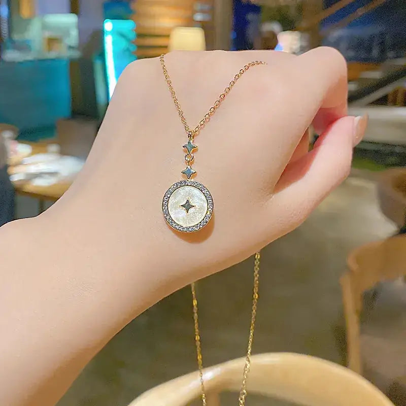Gold chain necklace with a circular pendant featuring an iridescent mother-of-pearl center and a diamond-studded border, accented by two small star-shaped charms above the main disc.
