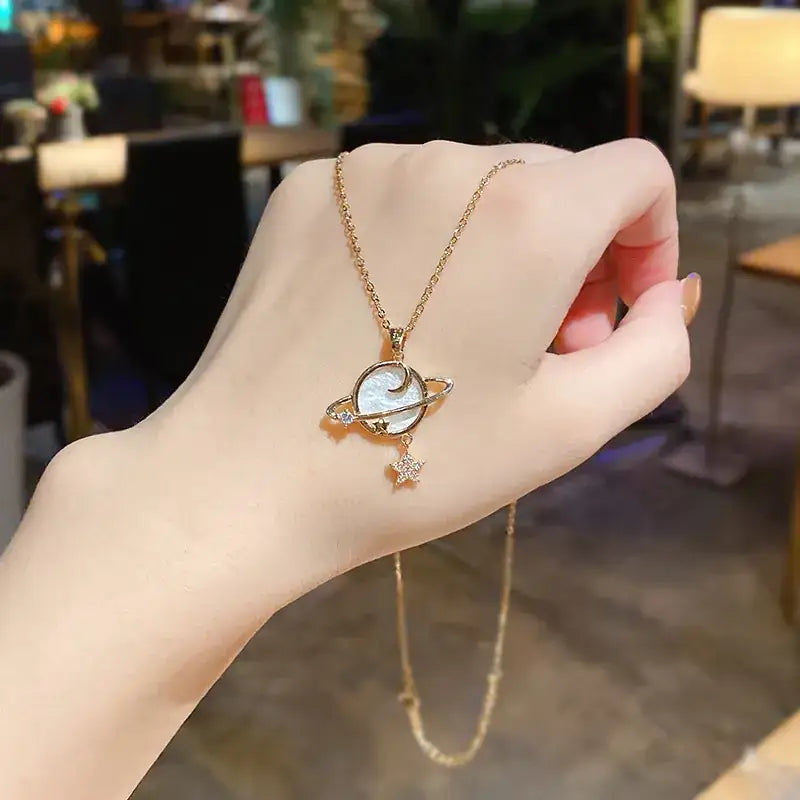 A delicate gold-toned necklace featuring a planet pendant with a white, iridescent center and sparkling diamond accents on its rings, accompanied by a small star charm.