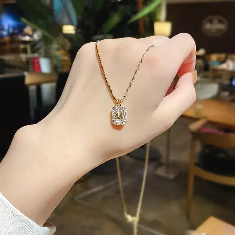 A gold-toned necklace with a rectangular pendant adorned with sparkling crystals and an initial ’m’ at its center.