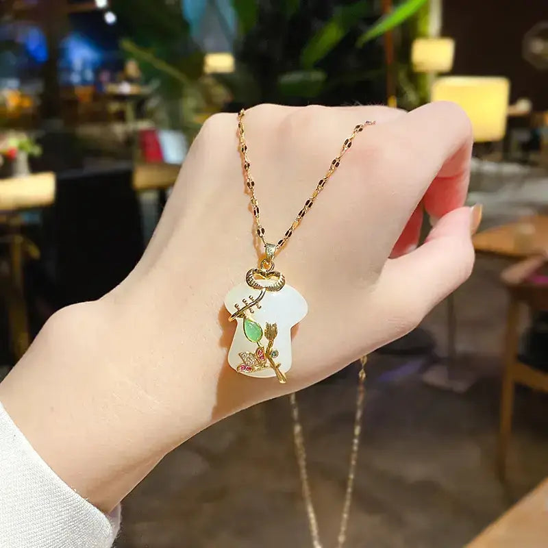 A delicate gold chain necklace featuring a white jade pendant shaped like an angelic figure, adorned with intricate golden details and small green gemstones.