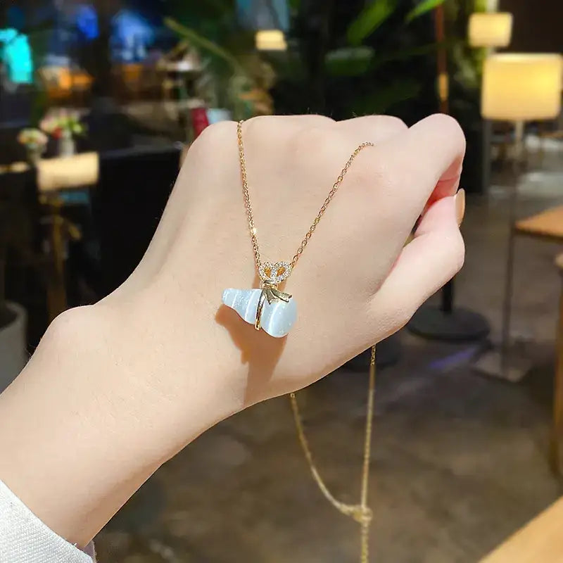 A delicate gold chain necklace featuring a light blue, translucent cat eye stone pendant shaped like a seashell with a small golden bow and diamond accents.