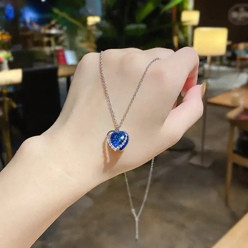 A delicate silver chain necklace featuring a heart-shaped pendant with a vibrant blue gemstone center and sparkling clear stones around its border.