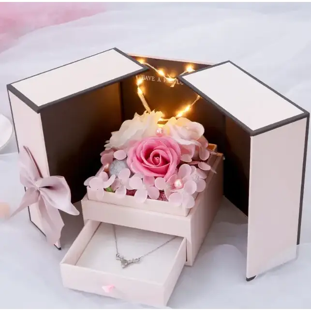 A delicate pink rose centerpiece surrounded by soft pastel flowers and sparkling embellishments, nestled inside a luxurious blush-colored gift box with an illuminated interior.