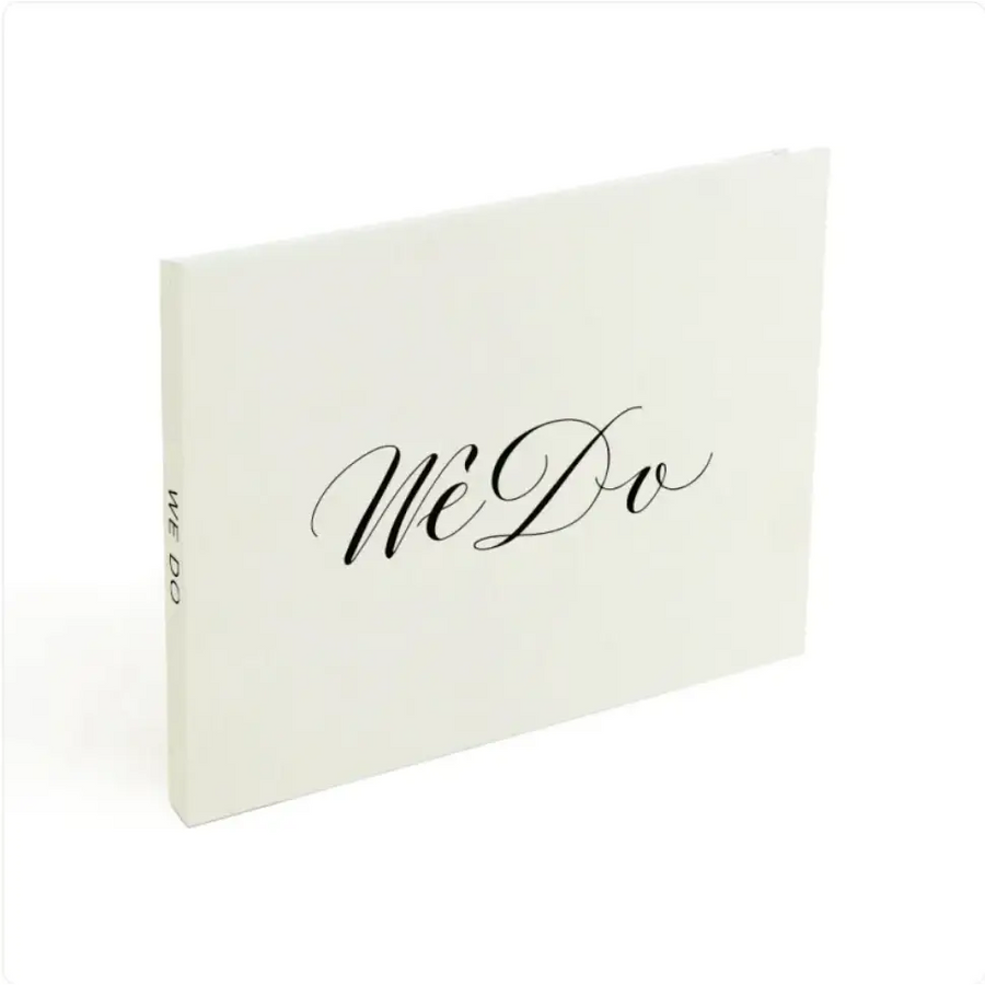 A minimalist white wedding guest book with elegant black cursive ’we do’ lettering on the cover.