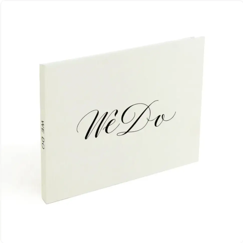 A minimalist white wedding guest book with elegant black cursive ’we do’ lettering on the cover.