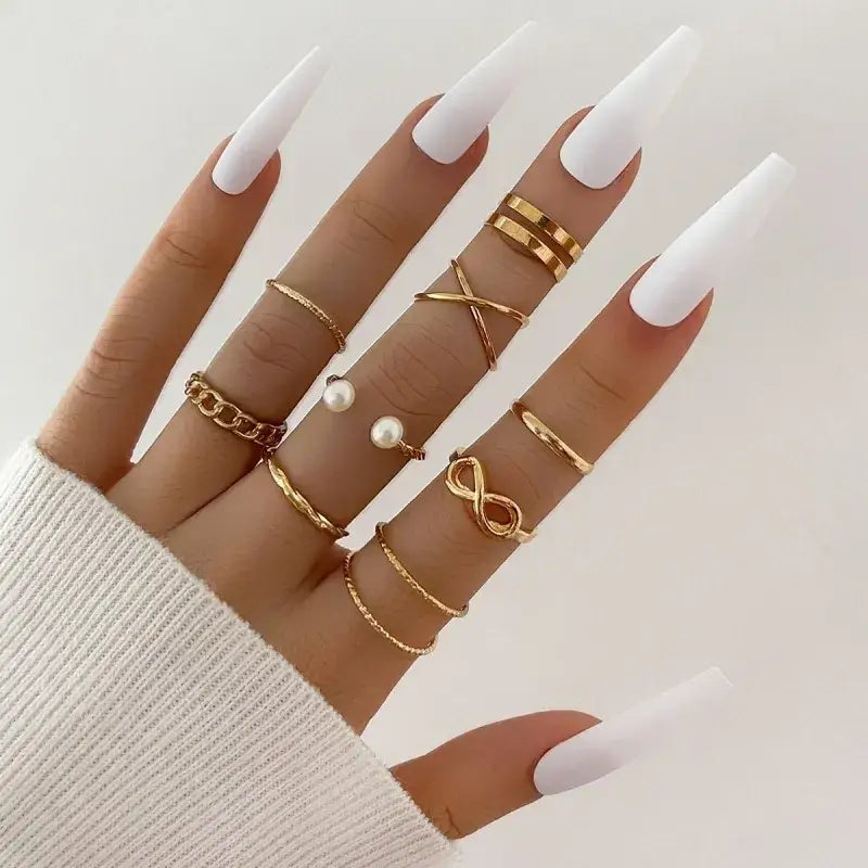 A hand adorned with multiple gold-toned rings featuring various designs including chains, infinity symbols, and pearl accents.