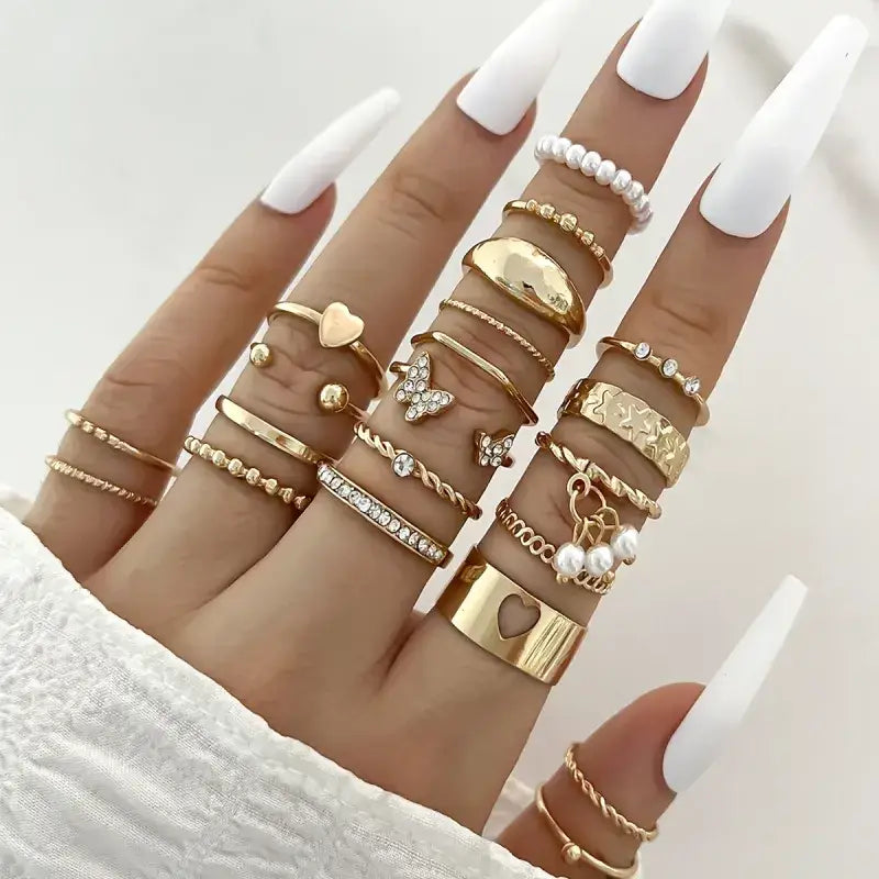 A hand adorned with multiple gold-toned rings featuring various designs including hearts, butterflies, pearls, and gemstones.