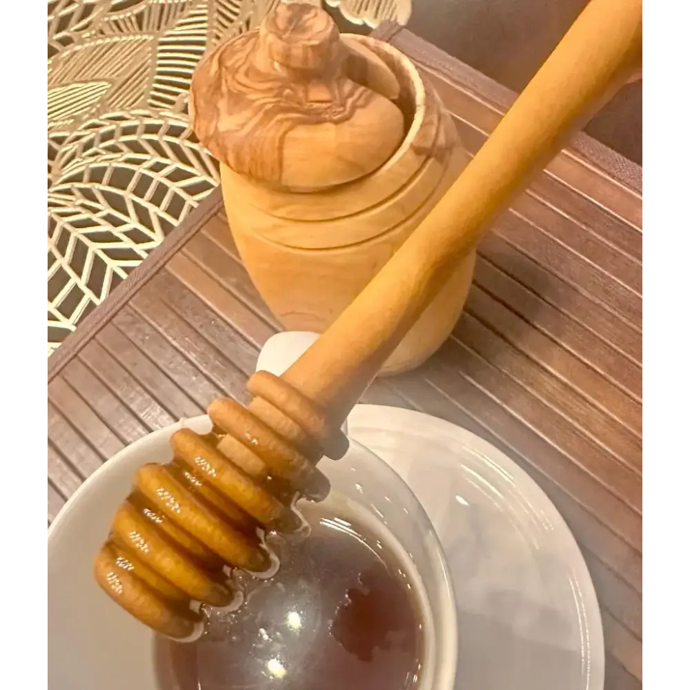 A wooden honey dipper with a smooth, light brown finish and rounded, grooved ends rests in a white bowl filled with golden amber honey.