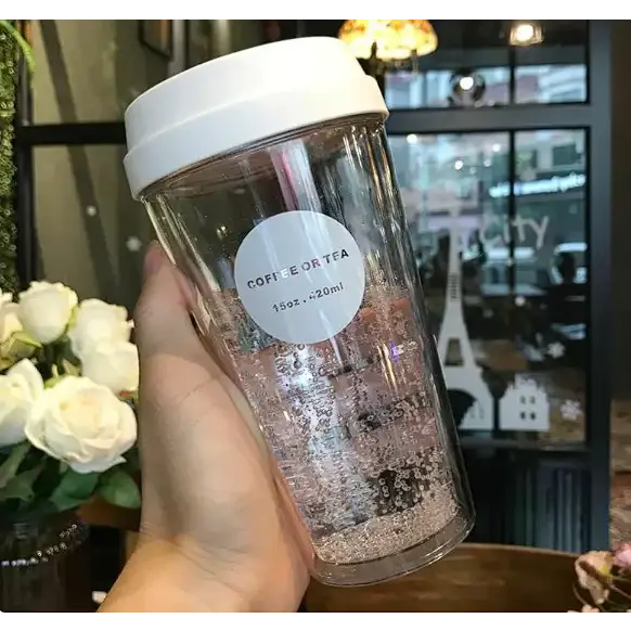 Clear plastic travel mug with a white lid and a circular label that reads ’coffee or tea 15oz.~20ml,’ filled with sparkling glittery liquid, held in someone’s hand against an indoor background featuring flowers and