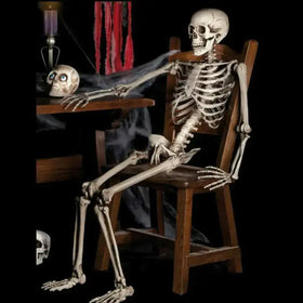 A detailed, off-white plastic skeleton figurine seated in a wooden chair with its arms resting on the table.