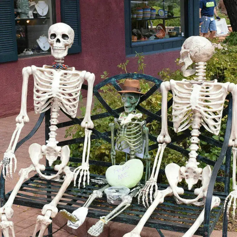 Three decorative skeleton figures, including a central green one wearing a brown hat and holding a heart-shaped sign that reads ’hug me,’ are seated on an outdoor bench.
