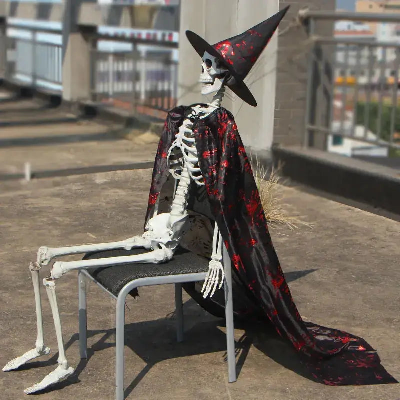 A white skeleton figure dressed in a black witch’s hat with red splatter patterns and draped in a matching black cape, seated on a metal chair.