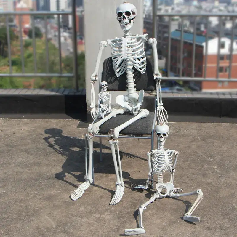 White plastic skeleton figurine seated on a chair with two smaller skeletons nearby.