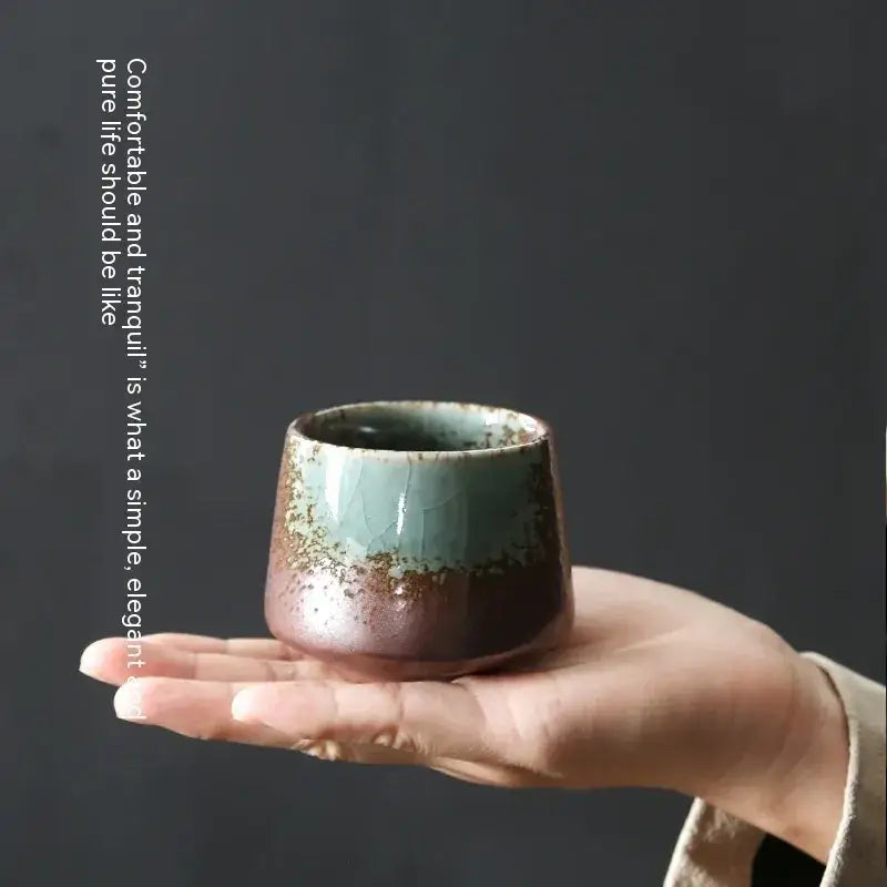 A hand holds a small, rustic ceramic tea cup with a gradient glaze transitioning from muted teal to earthy brown and speckled gold.
