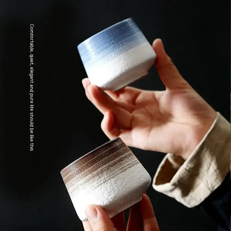 Two handcrafted ceramic tea cups, one with a soft blue-to-white gradient glaze and the other featuring earthy brown streaks over white.