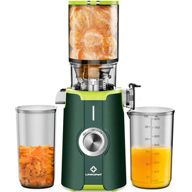 Green and silver linkohet juicer with transparent container filled with oranges, a glass measuring cup of orange juice to the right, and another glass jar containing chopped fruit on the left.