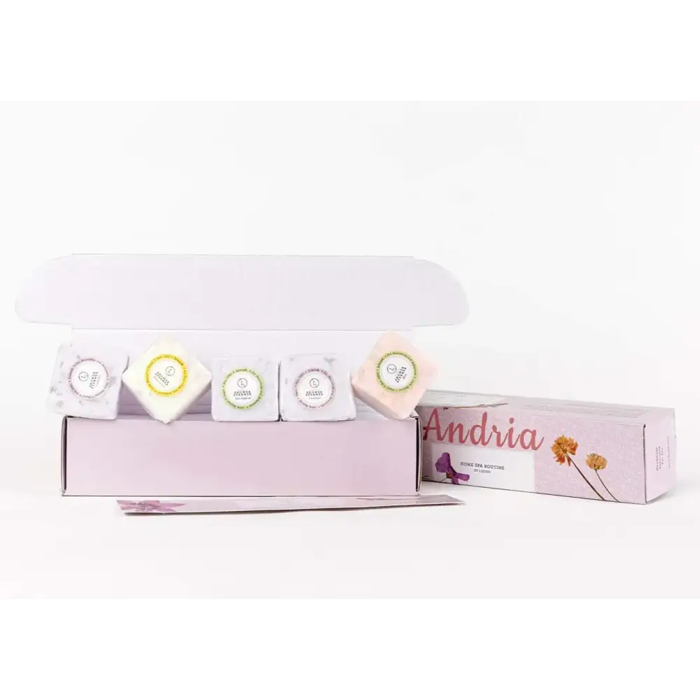 A lavender-colored gift box with a white lid, containing five square-shaped bath bombs in various pastel hues and designs.