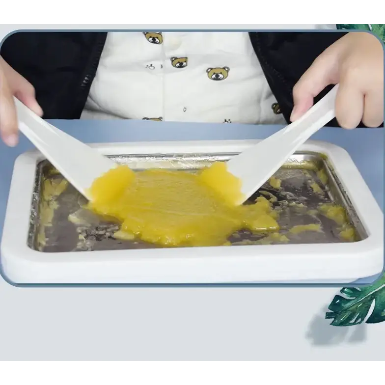 White rectangular ice cream making machine with two white plastic spatulas spreading yellow frozen mixture inside