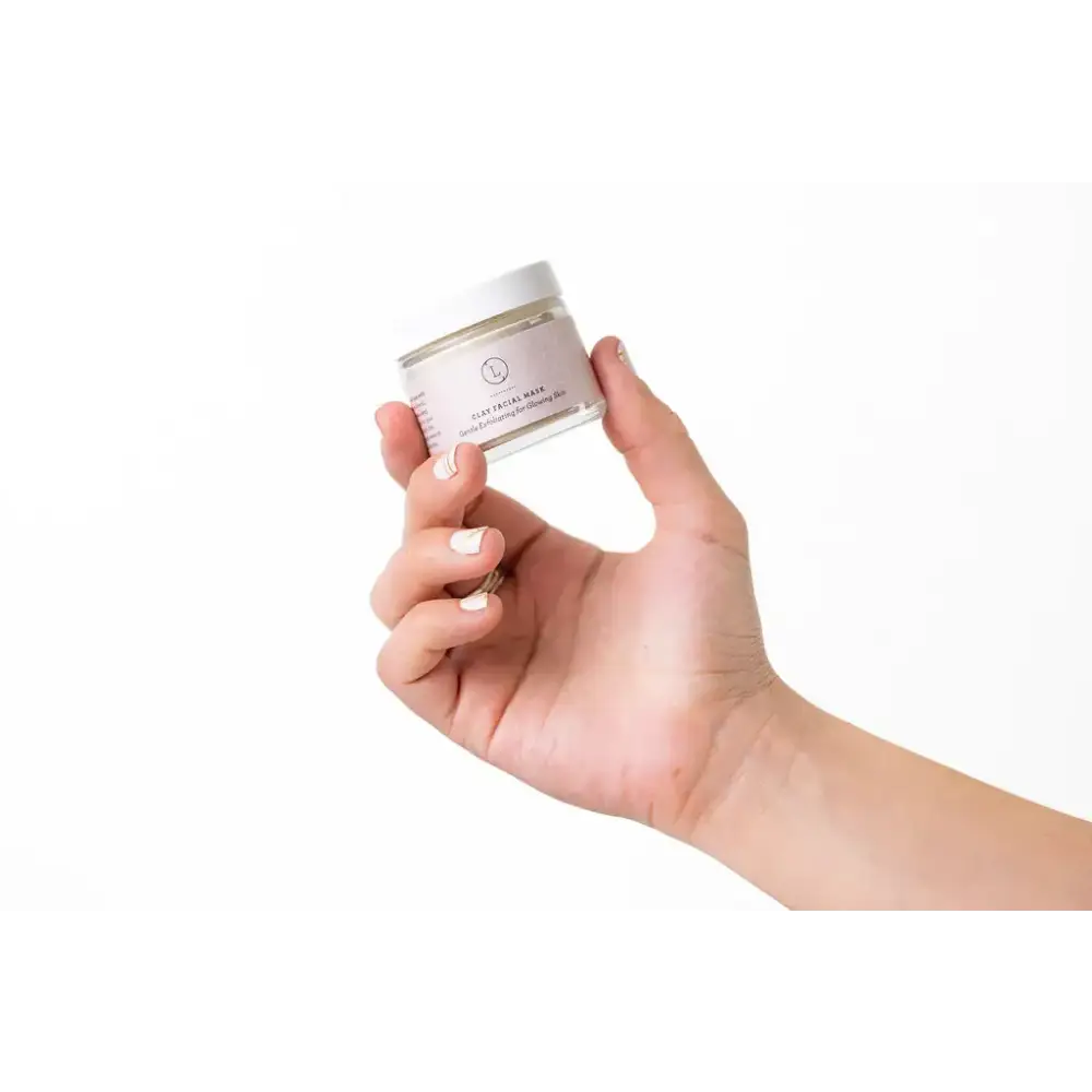 A clear glass jar with a white lid and minimalist label, holding a clay facial mask.