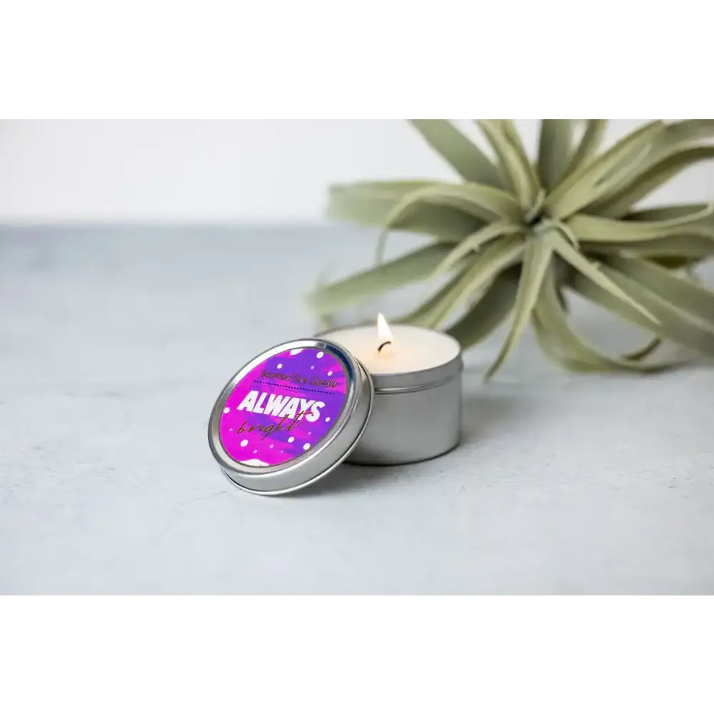 A lit, silver tin candle with a vibrant purple lid featuring the phrase ’always bright’ in white and gold lettering.