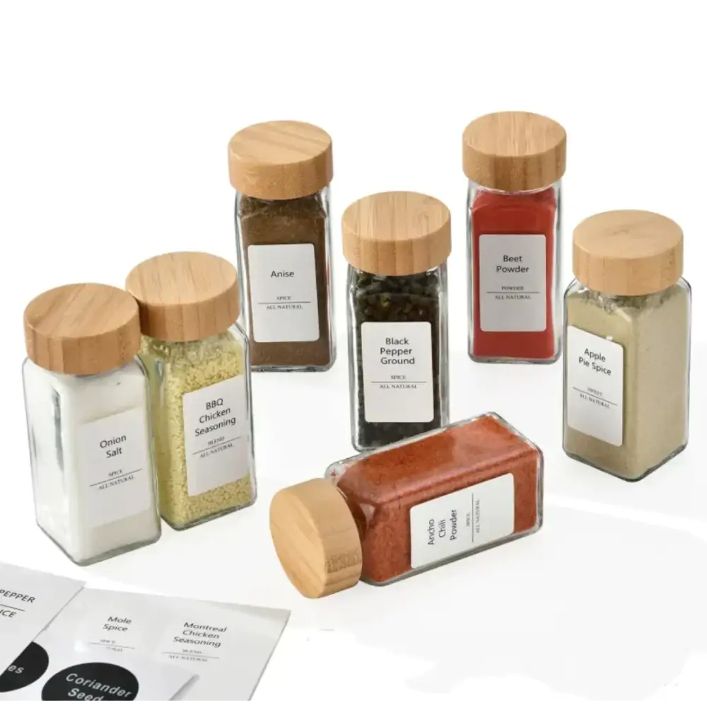 Set of clear glass spice jars with natural wood lids, each labeled with white stickers and containing different colored seasonings.
