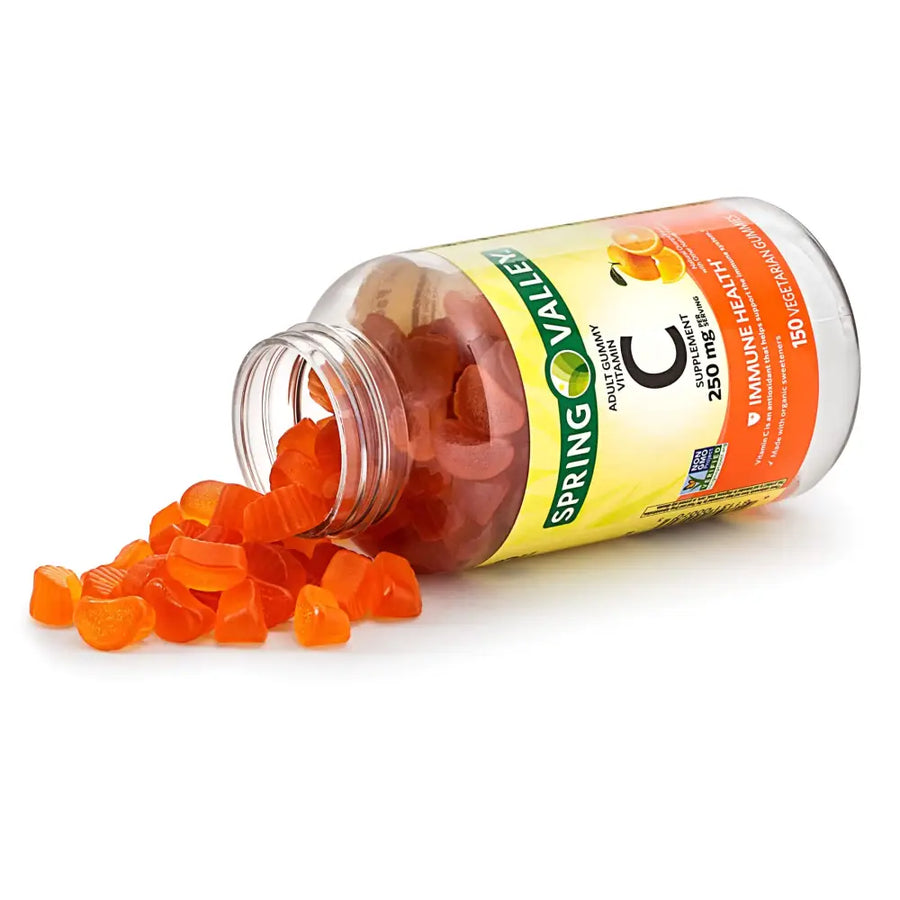 A clear glass bottle of spring valley adult gummy vitamin c, tipped over to spill bright orange citrus-shaped gummies onto a white surface.