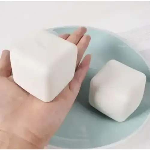 A smooth, off-white, cube-shaped bar of soap with rounded edges rests in a hand beside another similar bar on a light blue plate.