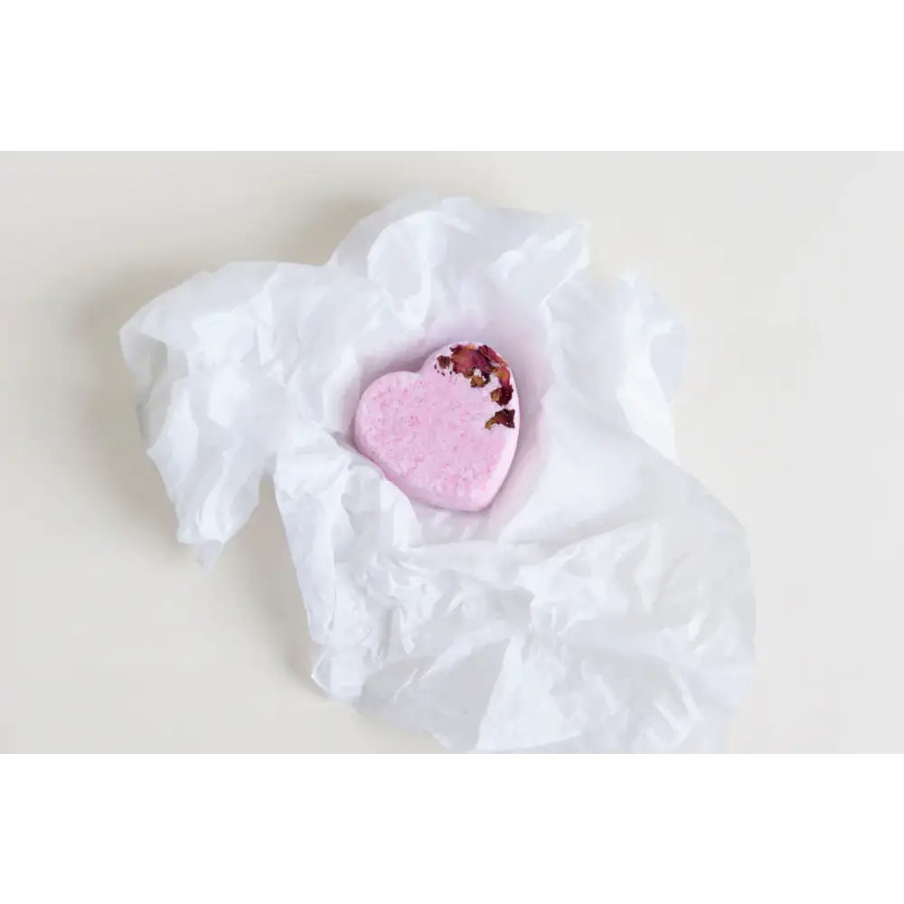 A pink heart-shaped bath bomb with dried rose petals embedded in its surface, nestled within crumpled white tissue paper.