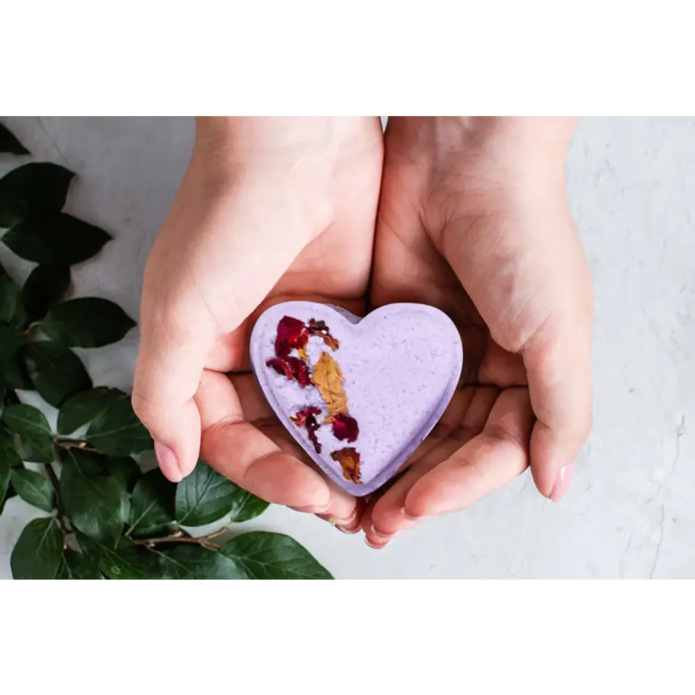 A lavender heart-shaped bath bomb with dried rose petals and gold leaf embedded in its surface.