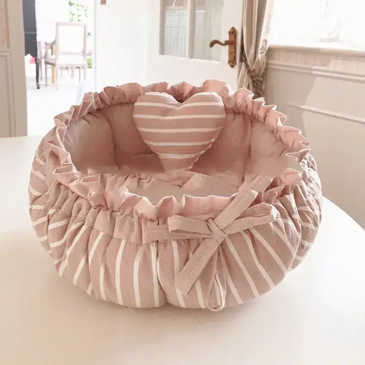 A soft pink and white striped fabric pet bed with a ruffled edge, featuring a matching heart-shaped pillow inside.