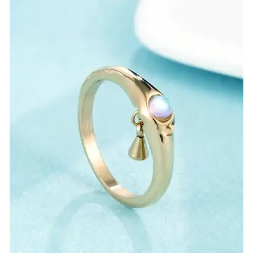 Gold-toned ring featuring a smooth, curved band with an embedded iridescent moonstone and a small dangling bell charm.