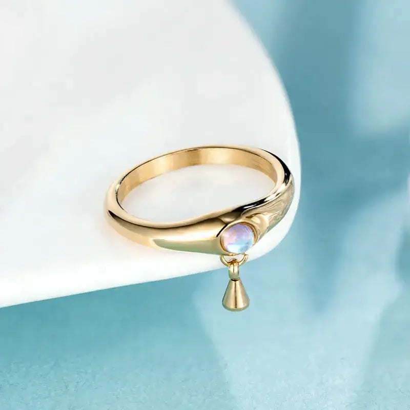 Gold-toned ring featuring a round, iridescent moonstone set in an elegant band with subtle engraved detailing and accented by a small teardrop-shaped pendant.
