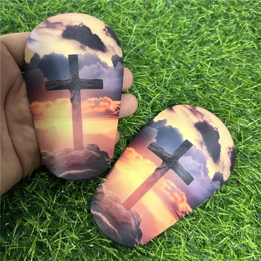 A pair of soft, oval-shaped baby footmuff covers featuring a printed design of a dark cross against a vibrant sunset sky with clouds.