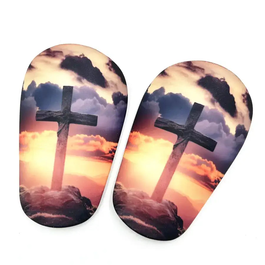 Baby booties featuring a printed design of a cross against a dramatic sunset sky with clouds, in vibrant orange and purple hues.