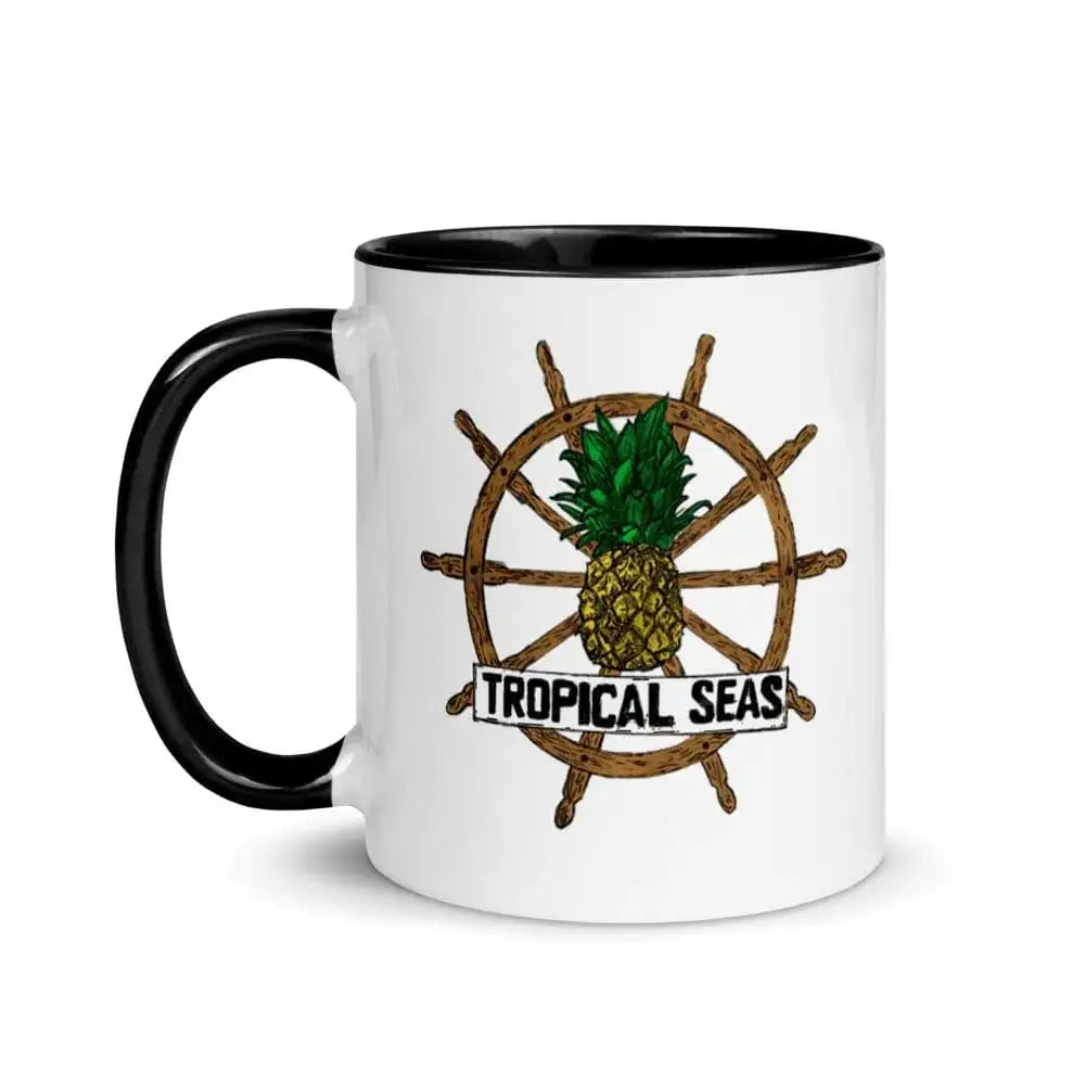 White ceramic mug with black rim and handle, featuring a tropical design of a pineapple centered on a wooden ship’s wheel with the text ’tropical seas’ below.
