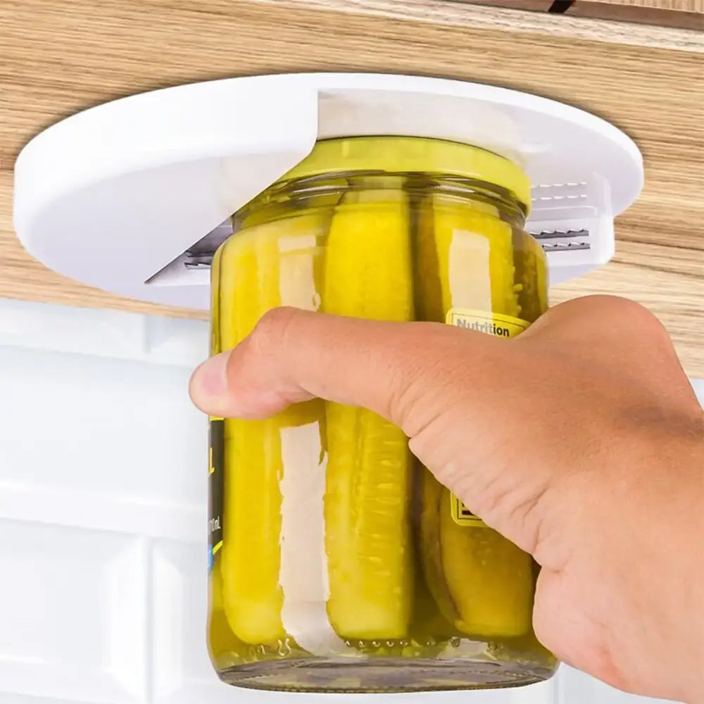 White plastic can opener mounted under a wooden countertop, gripping the lid of a glass jar filled with pickles