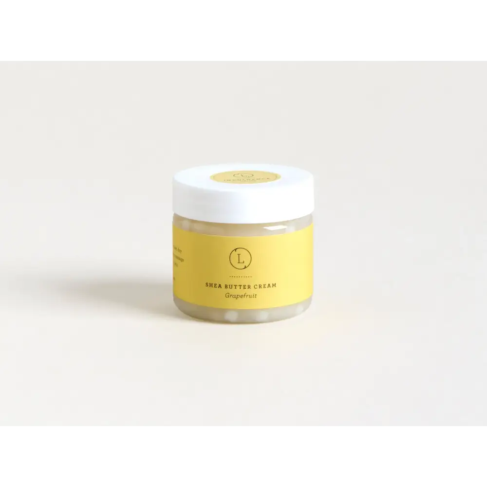 A clear glass jar with a white lid and yellow label, containing grapefruit-scented shea butter cream.