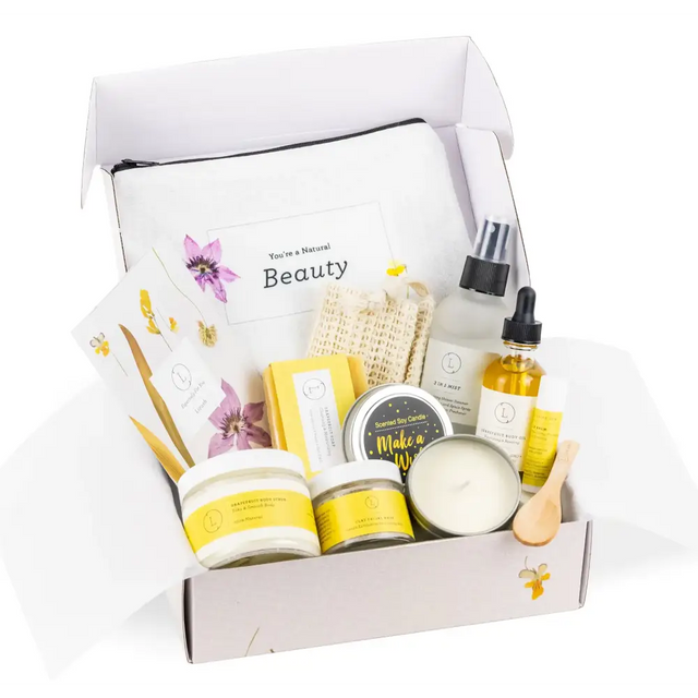 A white and yellow natural beauty gift set box containing candles, lotions, oils, a scrubber, and floral-themed accessories.