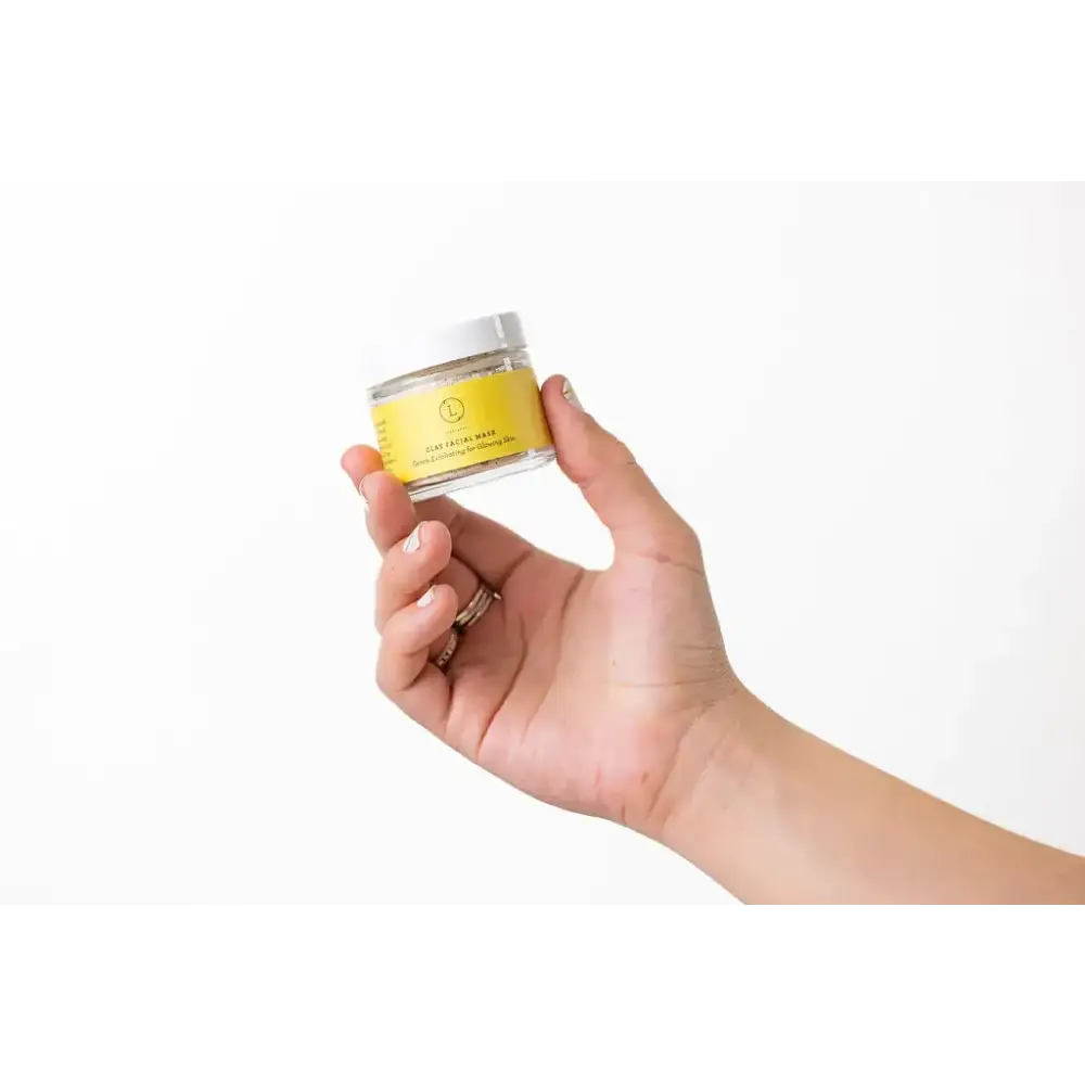 A clear glass jar with a white lid and a bright yellow label, containing a clay facial mask.