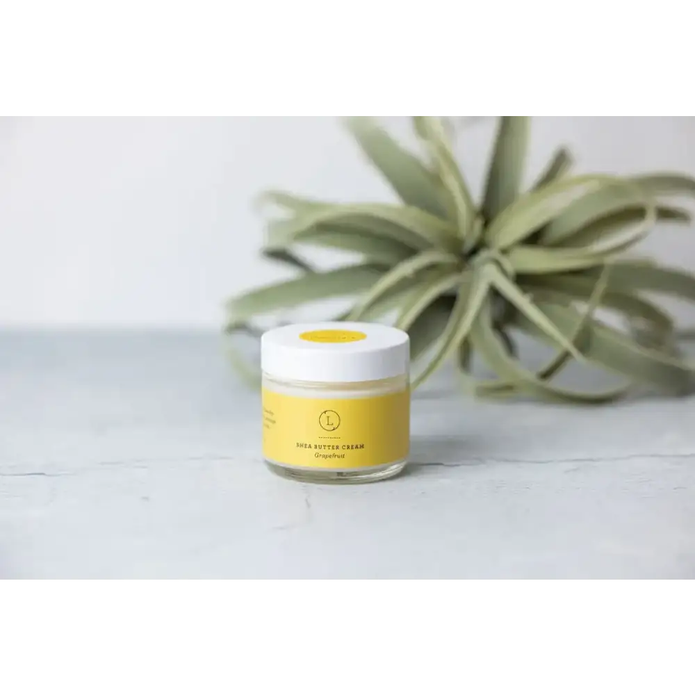 A clear glass jar with a white lid and yellow label, containing grapefruit-scented shea butter cream.