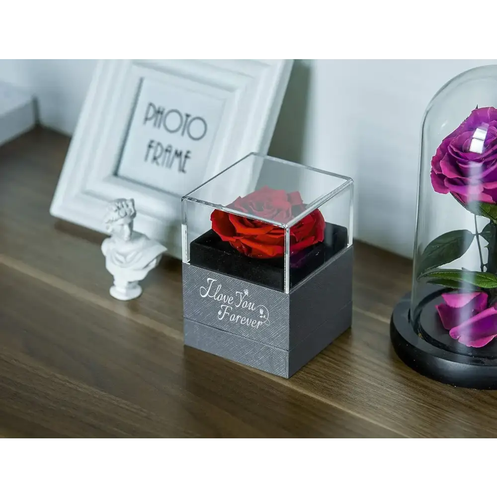 A vibrant red preserved rose nestled inside a clear acrylic cube with a textured gray base engraved with ’i love you forever.’