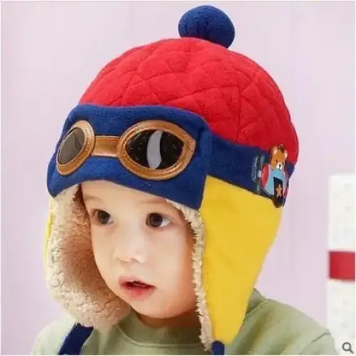 Red quilted winter hat with blue and yellow accents, faux fur trim, aviator goggles strap, and a navy pom-pom.