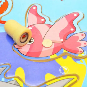 A yellow, cylindrical fishing lure with a red eye detail rests on the pink and white striped fish design.
