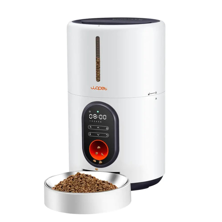 White cylindrical automatic pet feeder with black accents, digital display showing time and controls, orange brand logo, and a red feeding slot dispensing kibble into a silver bowl.